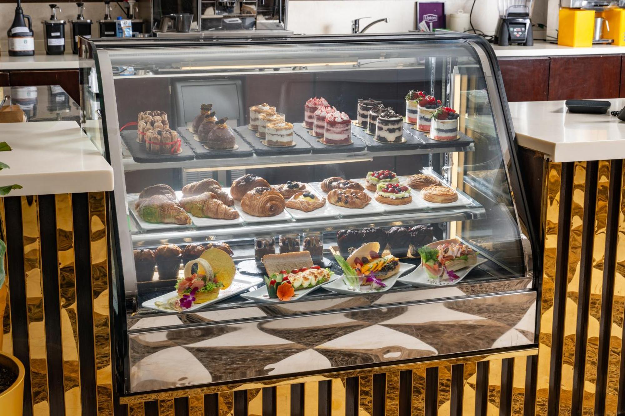 Al Aqeeq Madinah Hotel Medina Eksteriør billede The photo shows a display case filled with an assortment of desserts. There are various types of pastries, including cakes, tarts, and croissants, arranged neatly on different shelves. The desserts are visually appealing, with colorful decorations, f