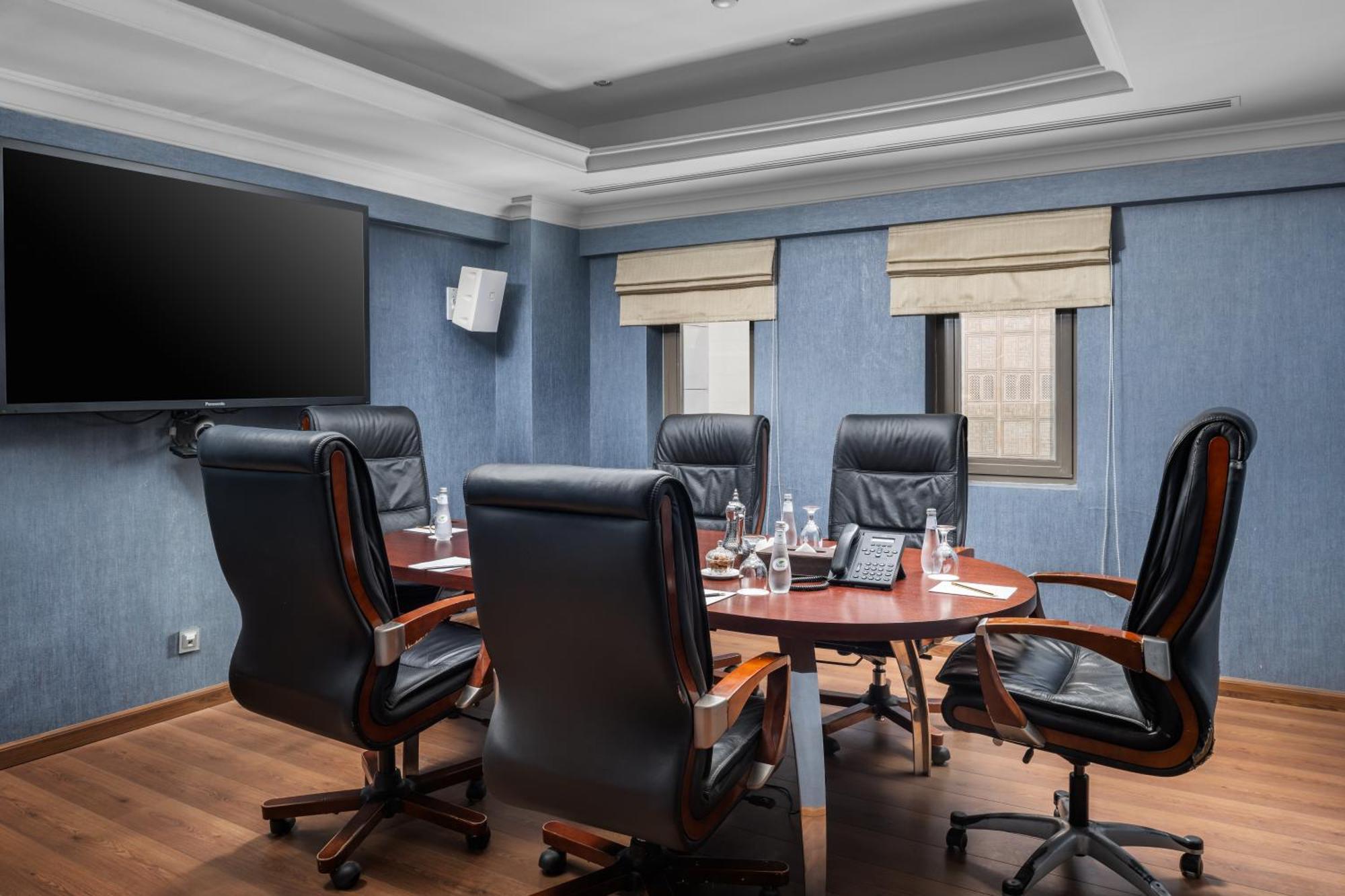 Al Aqeeq Madinah Hotel Medina Eksteriør billede The photo shows a modern conference room. It features a round wooden table surrounded by several black leather swivel chairs. The walls have a blue color scheme, and there is a large flat-screen TV mounted on one wall. The room has two windows with b