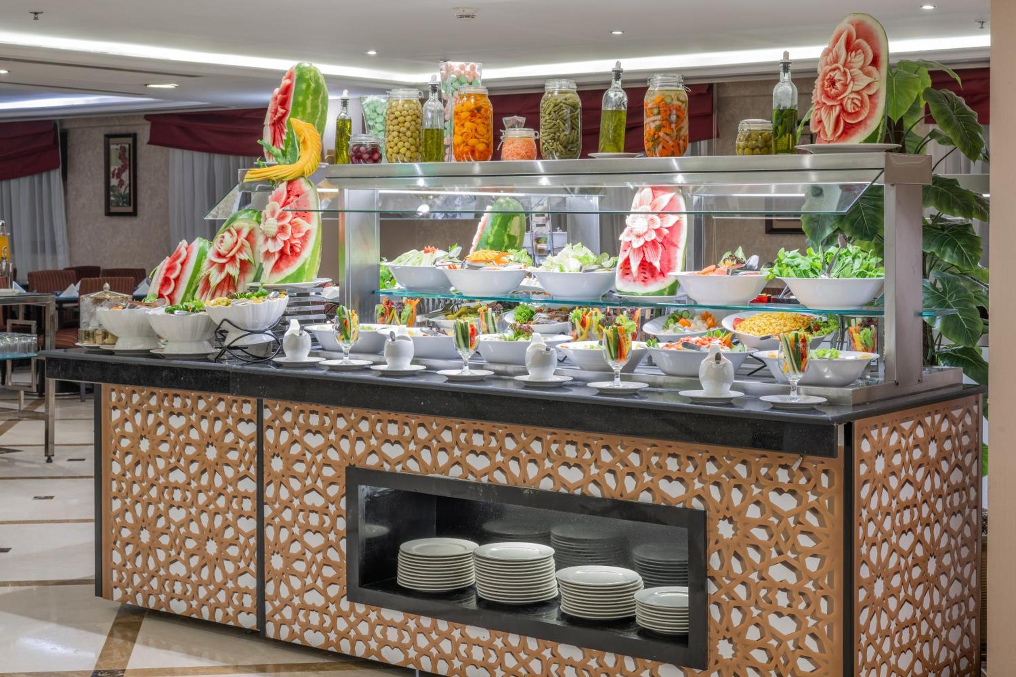 Al Aqeeq Madinah Hotel Medina Eksteriør billede The photo shows a buffet display that features an array of colorful food items. In the foreground, there are several large bowls filled with various salads and dishes. The presentation is visually appealing, with decorative watermelon carvings and an