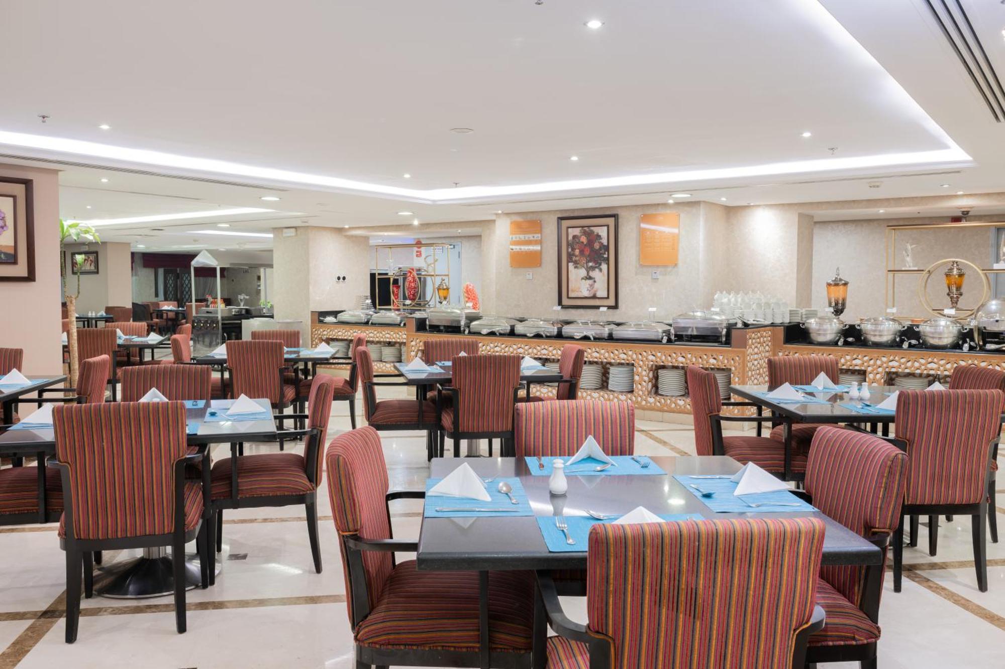 Al Aqeeq Madinah Hotel Medina Eksteriør billede The photo shows the interior of a restaurant. It features multiple tables arranged neatly, each with patterned chairs and set with blue and white table settings, including folded napkins. The lighting is bright and modern, creating a welcoming atmosp