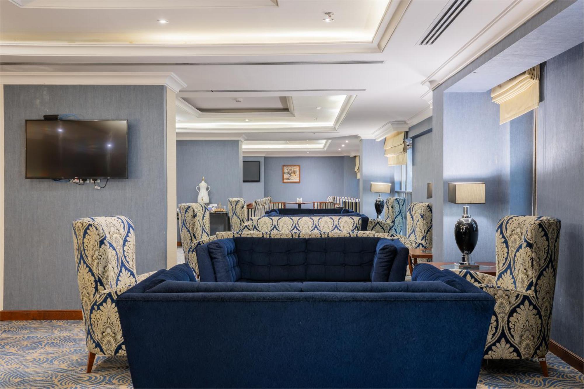 Al Aqeeq Madinah Hotel Medina Eksteriør billede The photo shows a stylish and modern lounge area. There are two patterned armchairs with blue and cream designs, positioned next to a deep blue couch. The room has a light blue wall with a flat-screen TV mounted on one side. Soft lighting from stylis