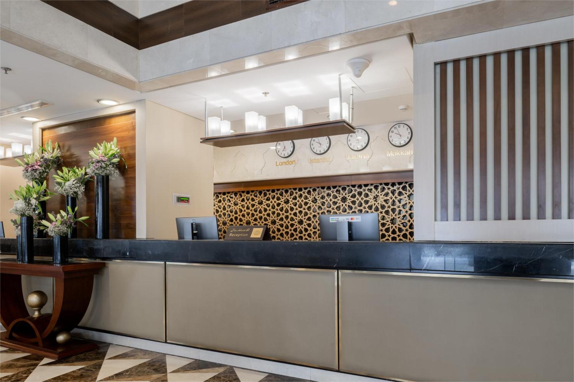 Al Aqeeq Madinah Hotel Medina Eksteriør billede The photo shows the interior of a hotel reception area. The reception desk is designed with a sleek, modern look featuring a dark countertop and a light-colored front. Above the desk, there are several clocks displaying different times, suggesting an