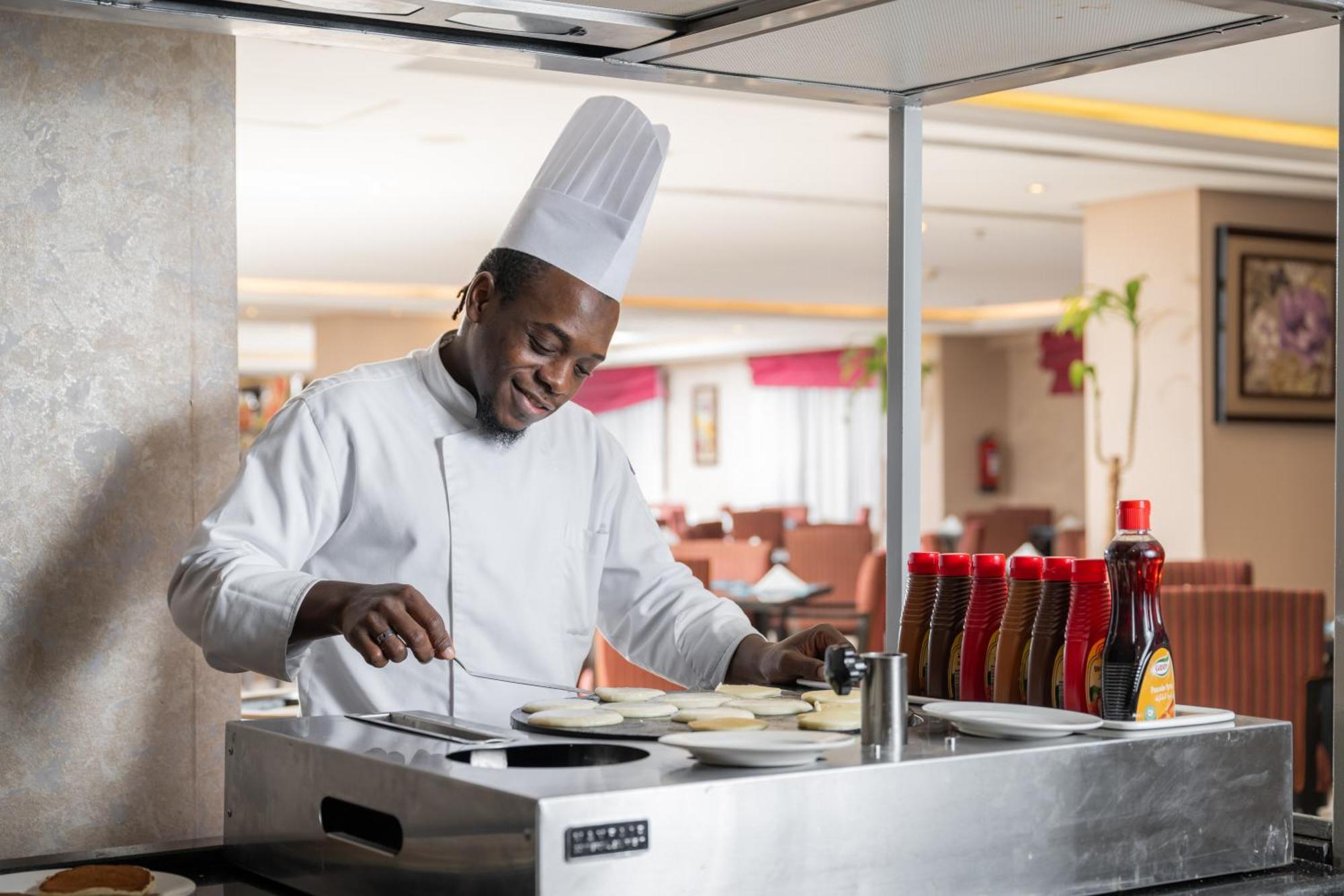 Al Aqeeq Madinah Hotel Medina Eksteriør billede The photo shows a chef in a professional kitchen setting. He is wearing a traditional white chef's uniform and a tall white chef's hat, and he appears to be smiling while cooking. The chef is standing at a stainless steel cooking station, where he is