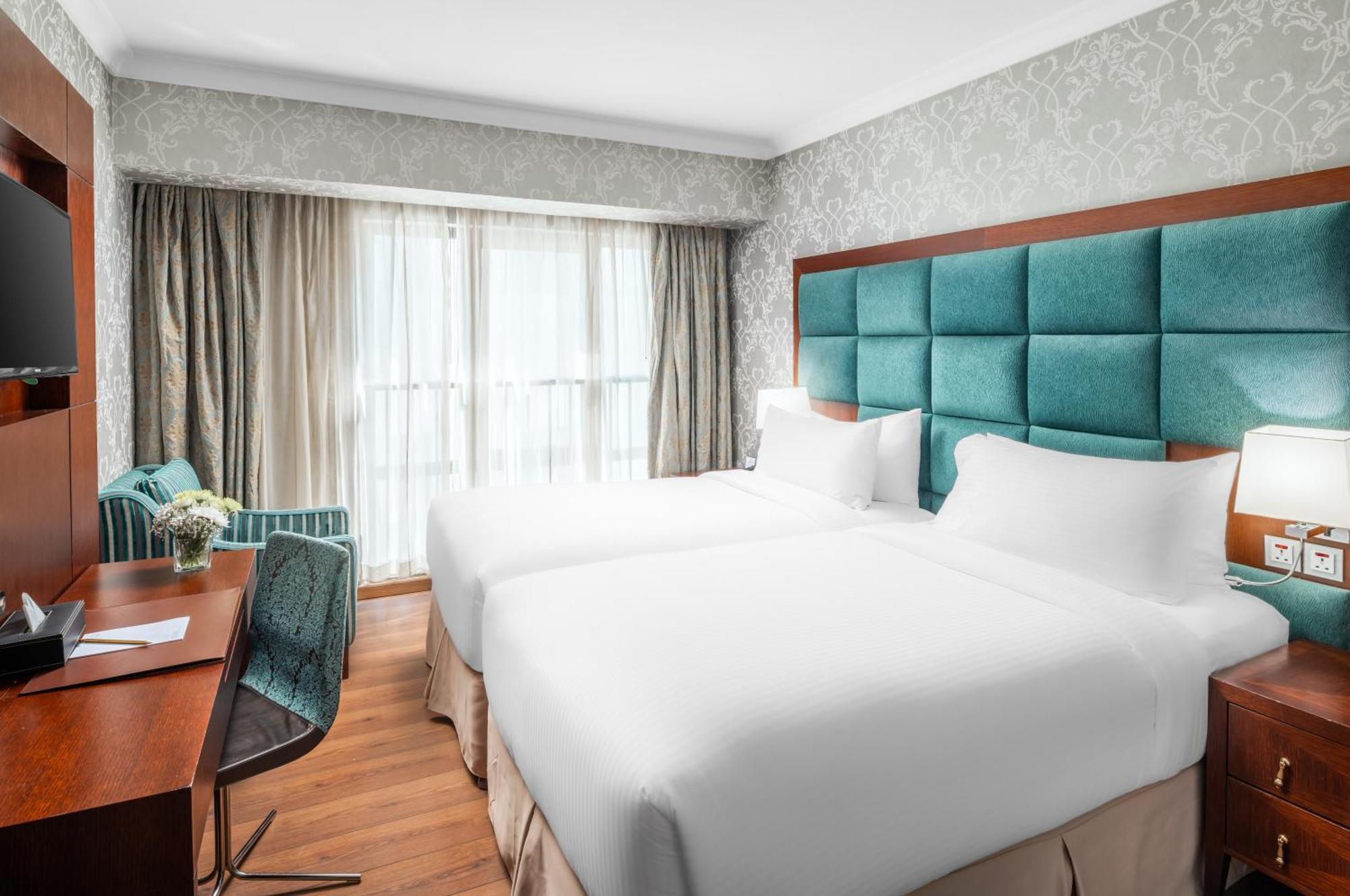 Al Aqeeq Madinah Hotel Medina Eksteriør billede The photo shows a modern hotel room featuring two single beds with white bedding. The headboard is upholstered in a teal fabric. There is a wooden desk and a chair near a window that is covered with sheer curtains and drapes. A cozy armchair in a mat