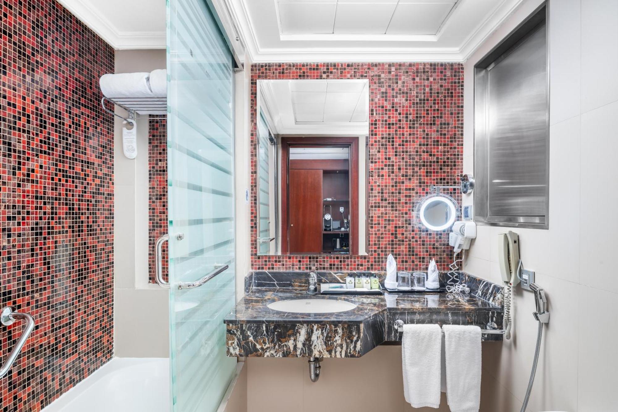Al Aqeeq Madinah Hotel Medina Eksteriør billede The photo shows a modern bathroom featuring a mix of design elements. The walls have a vibrant red mosaic tile pattern, adding a bold touch to the space. There is a glass shower enclosure, and next to it, a sleek countertop with a dark, marbled surfa