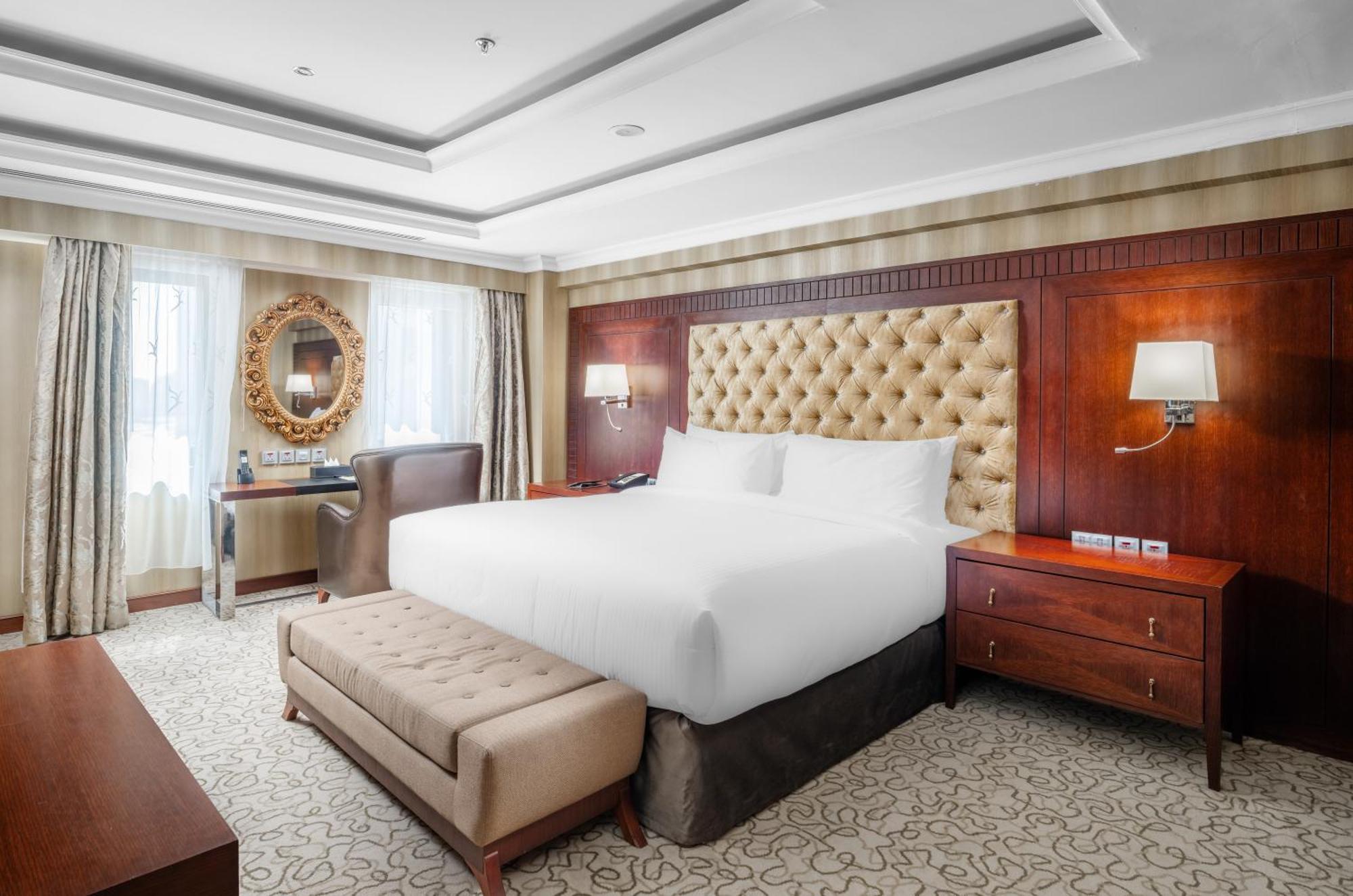 Al Aqeeq Madinah Hotel Medina Eksteriør billede The photo shows a well-furnished hotel room featuring a large bed with a tufted headboard. The room has a warm color palette with wooden accents and soft lighting. There are two windows with curtains that allow natural light to fill the space. Alongs
