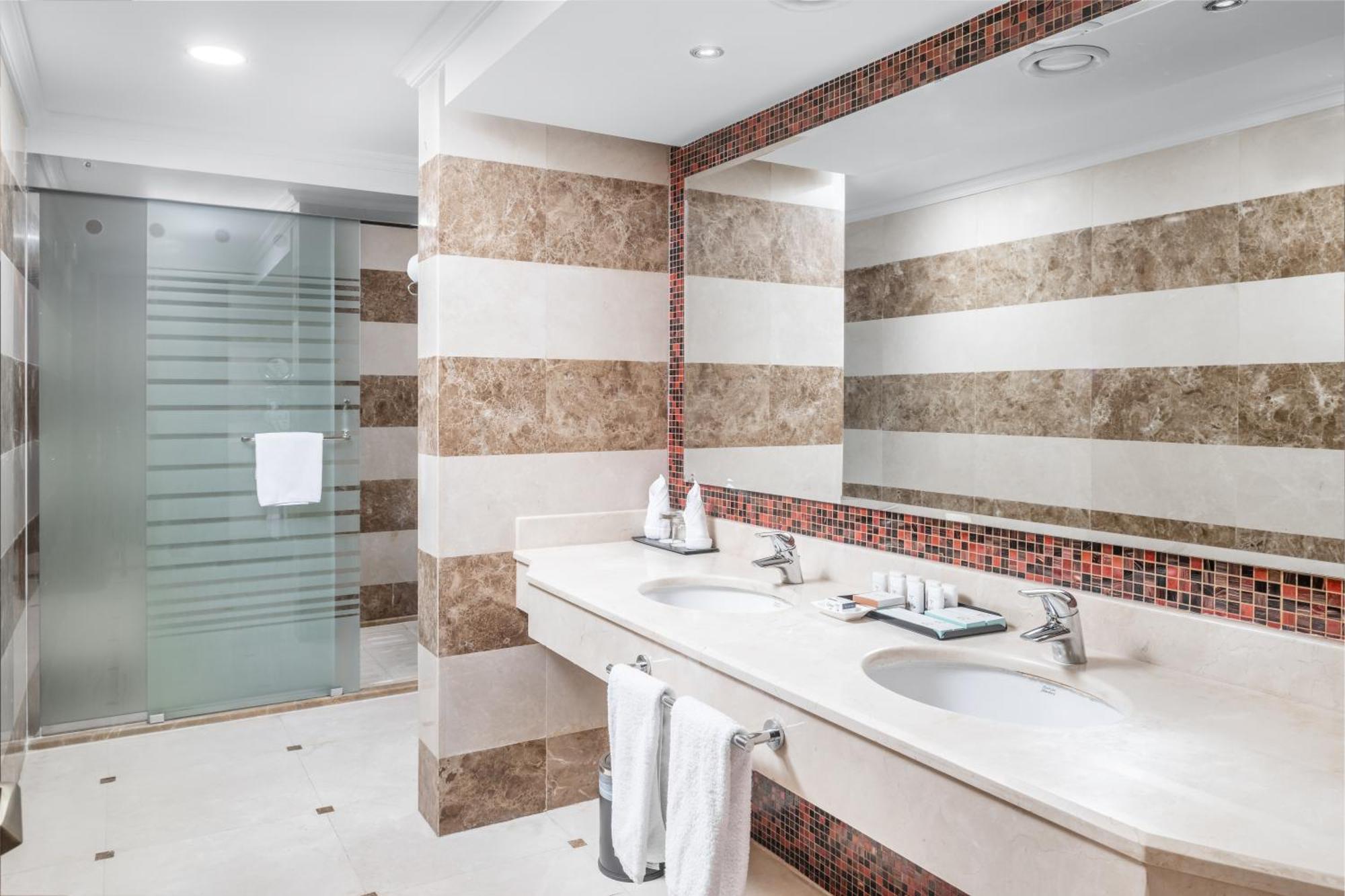 Al Aqeeq Madinah Hotel Medina Eksteriør billede The photo shows a modern bathroom with an elegant design. It features a large double sink with a smooth countertop, accompanied by faucets. The walls are adorned with alternating horizontal stripes of light and dark materials, creating a stylish look