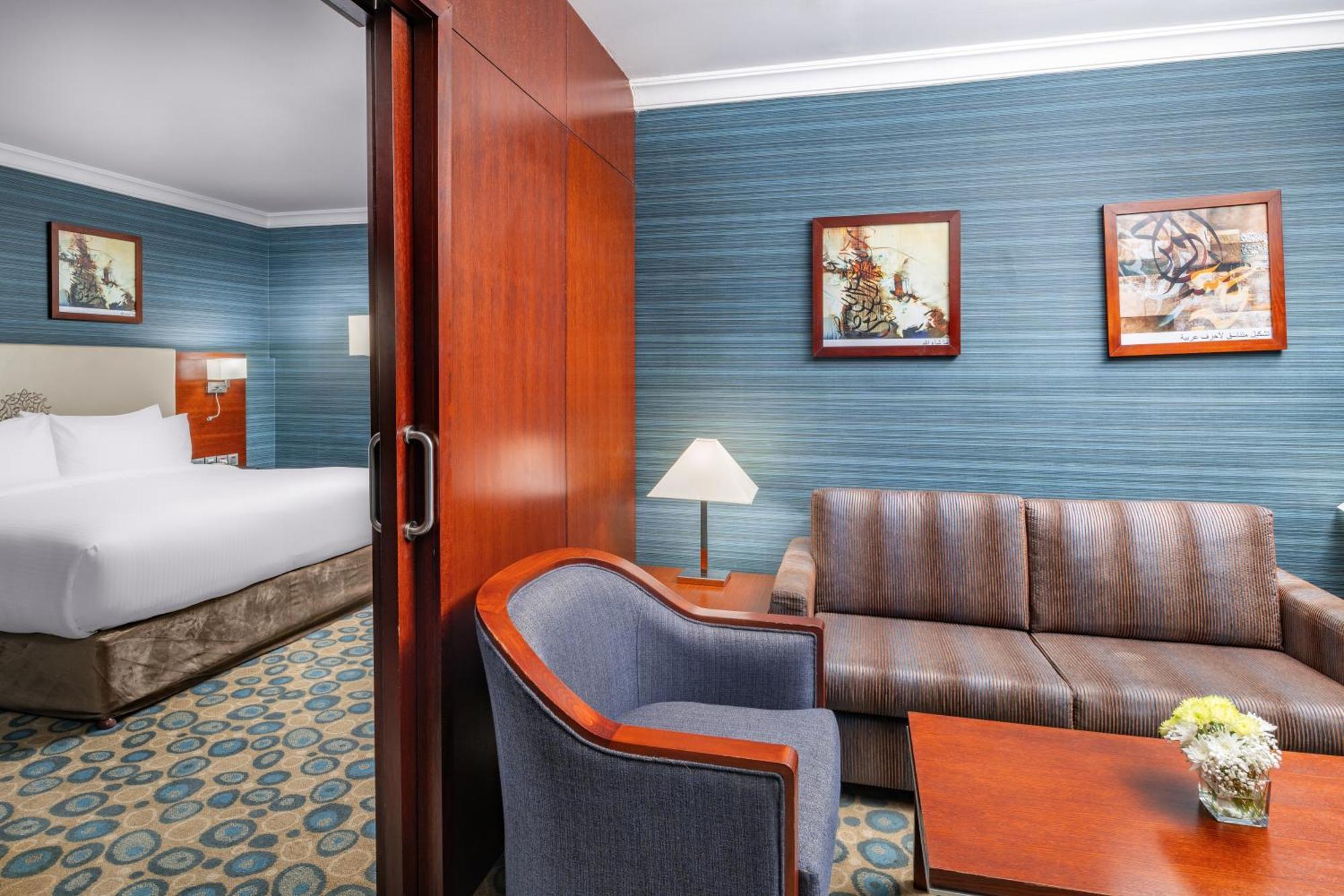 Al Aqeeq Madinah Hotel Medina Eksteriør billede The photo shows a well-furnished hotel room featuring a living area and a bedroom. To the left, there is a bed visible through an open doorway. The living area includes a gray upholstered chair and a brown couch, both arranged around a small table. A