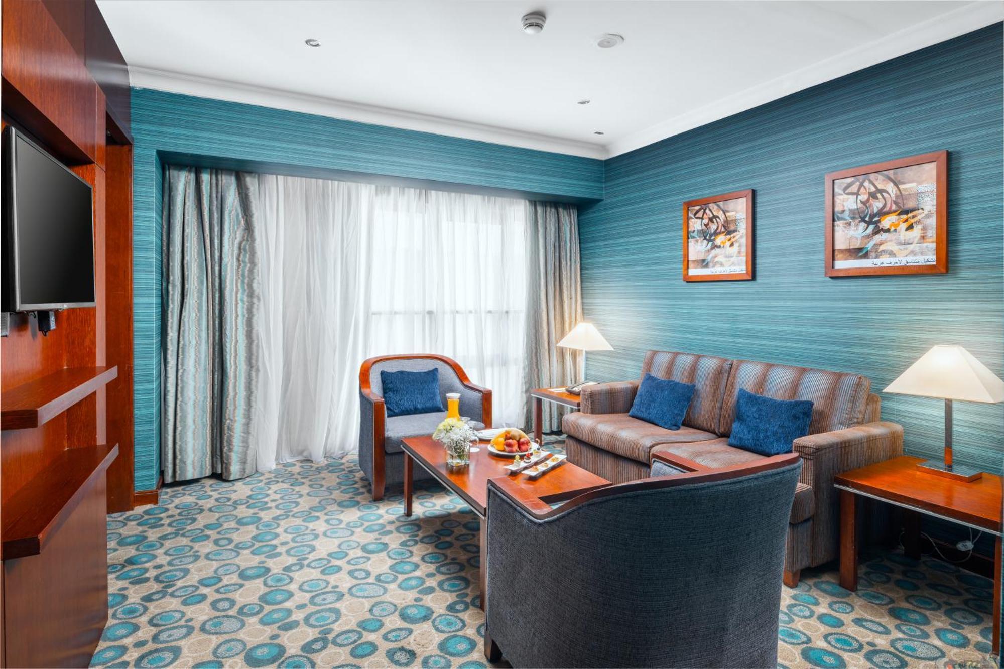 Al Aqeeq Madinah Hotel Medina Eksteriør billede The photo shows a cozy and modern hotel room featuring a sitting area. The walls are painted a calming teal color, complemented by a patterned carpet in shades of blue and beige. There is a comfortable sofa with cushions, two armchairs, and a wooden 