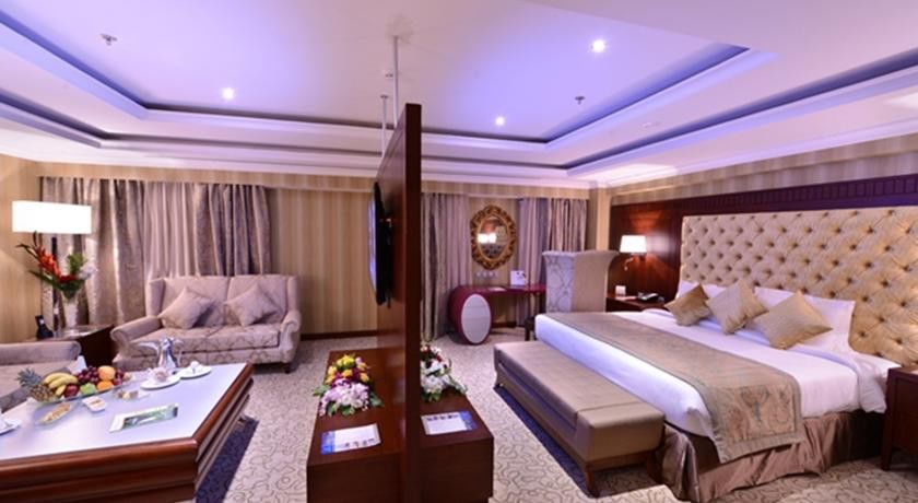 Al Aqeeq Madinah Hotel Medina Eksteriør billede The image shows a luxurious hotel room. It features a large bed with a tufted headboard, flanked by nightstands with lamps. There's a seating area with a plush sofa and a coffee table. A decorative partition separates the bed from the living area. Th