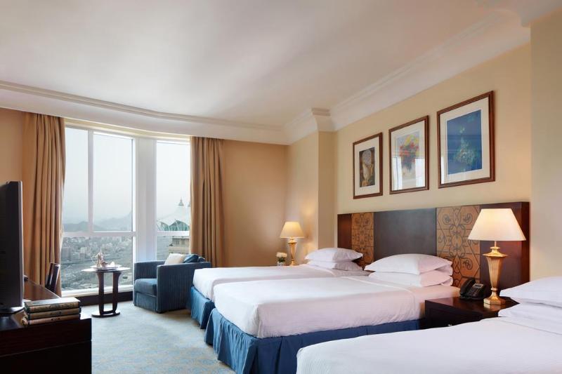 Al Aqeeq Madinah Hotel Medina Eksteriør billede The photo shows a hotel room featuring three beds arranged neatly. The room has a warm, inviting ambiance with beige walls and a soft carpet. Large windows allow natural light to fill the space, offering a view outside. There are curtains in a light 