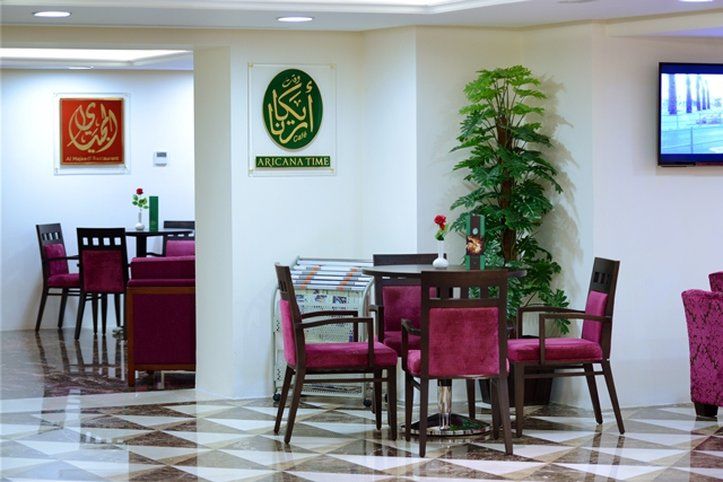 Al Aqeeq Madinah Hotel Medina Eksteriør billede The photo shows an interior space, likely a dining or lounge area. There are several tables with four chairs each, featuring a modern design with dark wood and plush maroon upholstery. On the walls, there are decorative signs, one of which has Arabic